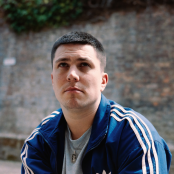 Aaron rowe sitting in front of brick wall wearing Adidas tracksuit